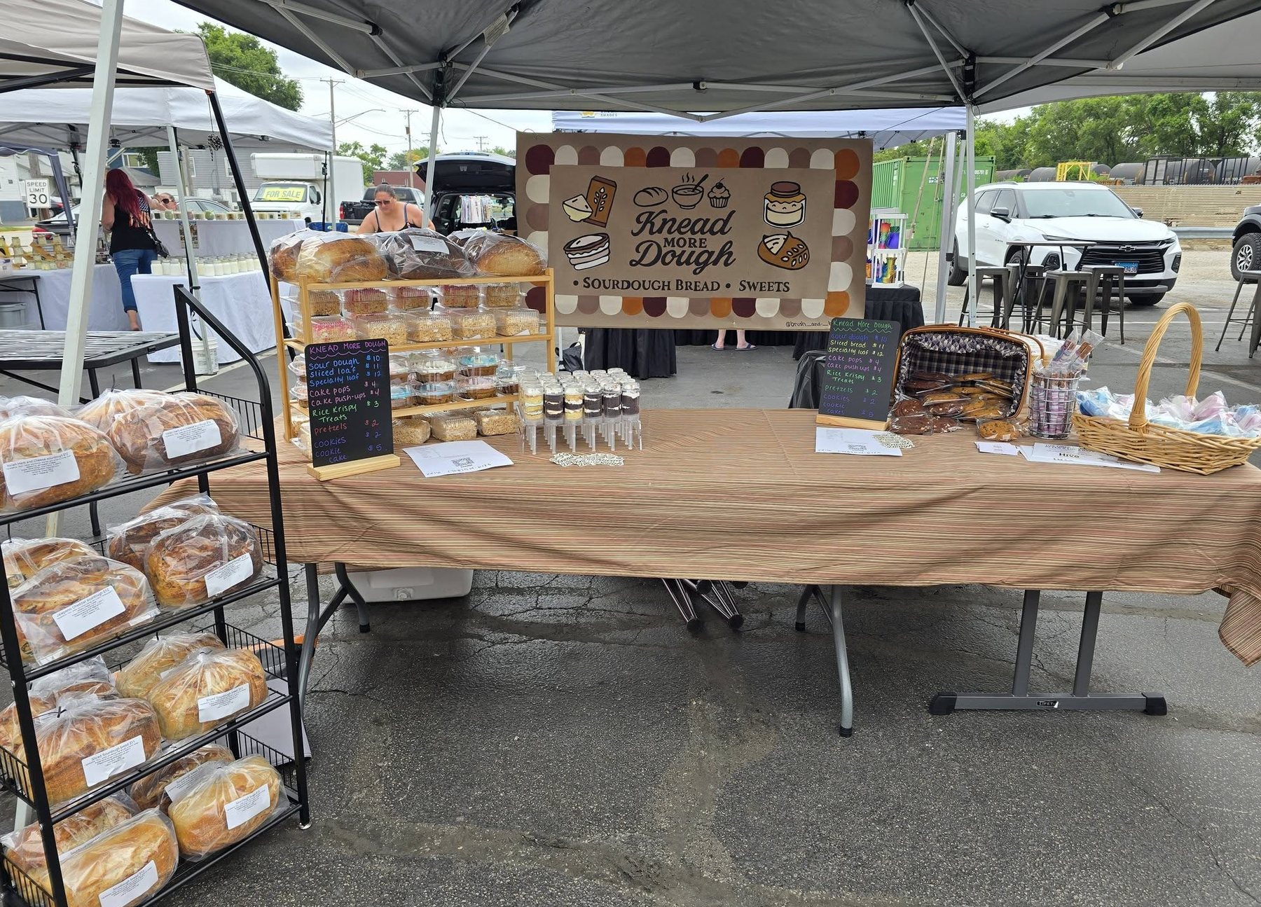 The Knead More Dough market booth with the painted banner, racks of bagged sourdough and a table of sweets